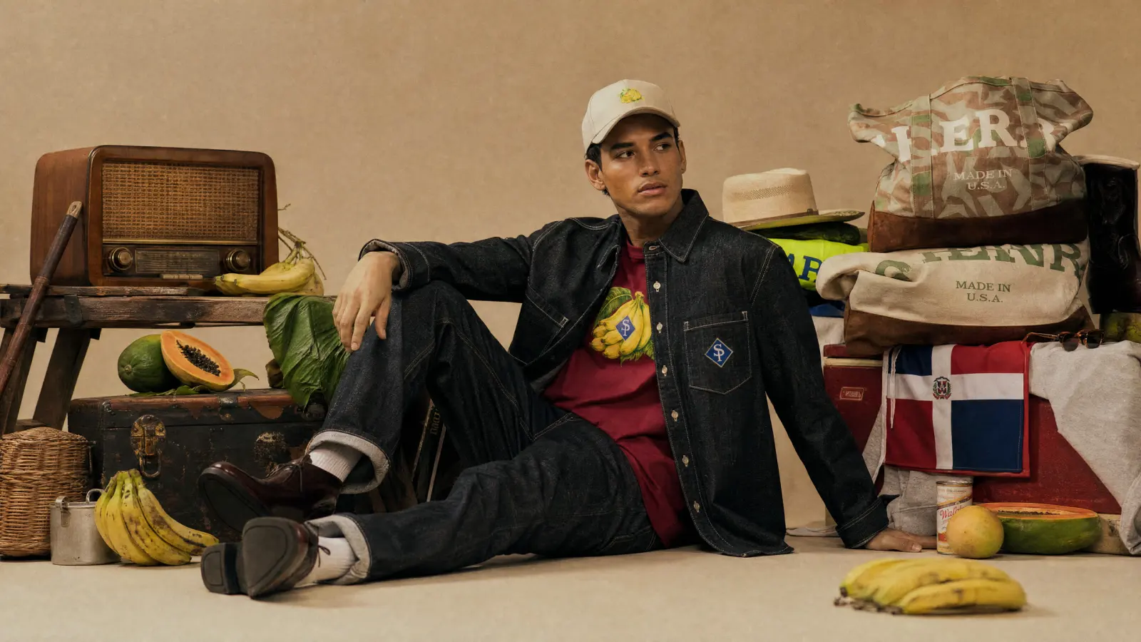Chaqueta vaquera sobre camiseta vino Guineo Niño y gorra crema, sentado en el estudio entre fruta y sacos
